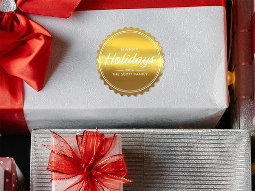 A gold foil cardstock Happy Holidays gift tag is shown on a Christmas present. The present is wrapped in white paper and has a red bow and ribbon around it. It has other Christmas presents surrounding it.