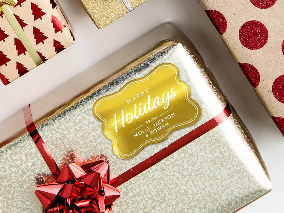 A gold foil cardstock Happy Holidays gift tag is shown on a Christmas present. The present is wrapped in gold paper and has a red bow and ribbon around it. The background of the picture is a white surface.