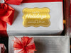 A gold foil cardstock Happy Holidays gift tag is shown on a Christmas present. The present is wrapped in white paper and has a red bow and ribbon around it. It has other Christmas presents surrounding it.