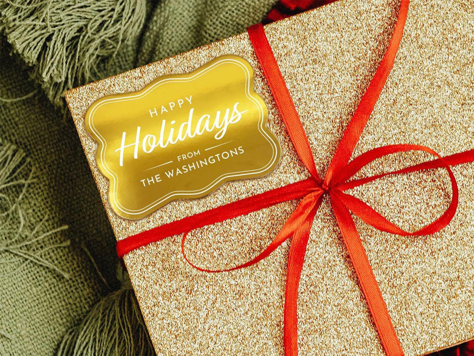 A gold foil cardstock Happy Holidays gift tag is shown on a glitter wrapped box. The box also has a red ribbon on it. The box is seen on a green blanket surface.