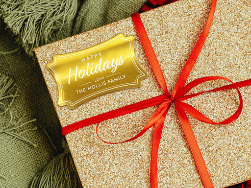 A gold foil cardstock Happy Holidays gift tag is shown on a glitter wrapped box. The box also has a red ribbon on it. The box is seen on a green blanket surface.