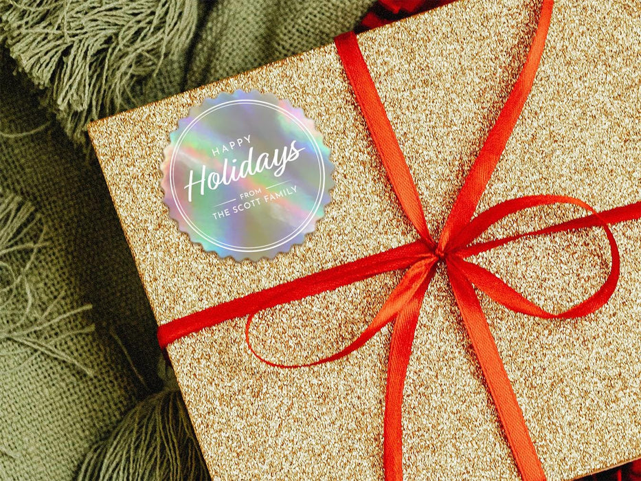 A rainbow cardstock Happy Holidays gift tag is shown on a glitter wrapped box. The box also has a red ribbon on it. The box is seen on a green blanket surface.