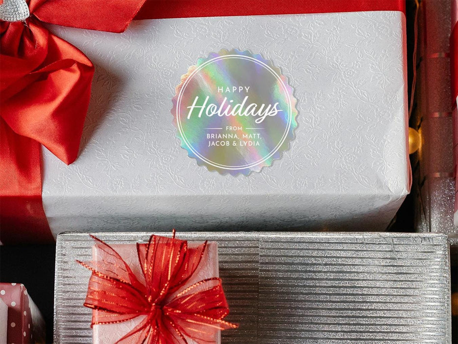 A rainbow cardstock Happy Holidays gift tag is shown on a Christmas present. The present is wrapped in white paper and has a red bow and ribbon around it. It has other Christmas presents surrounding it.