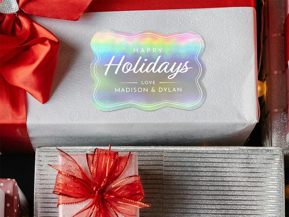 A rainbow cardstock Happy Holidays gift tag is shown on a Christmas present. The present is wrapped in white paper and has a red bow and ribbon around it. It has other Christmas presents surrounding it.