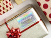 A rainbow cardstock Happy Holidays gift tag is shown on a Christmas present. The present is wrapped in gold paper and has a red bow and ribbon around it. The background of the picture is a white surface.