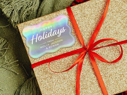 A rainbow cardstock Happy Holidays gift tag is shown on a glitter wrapped box. The box also has a red ribbon on it. The box is seen on a green blanket surface.