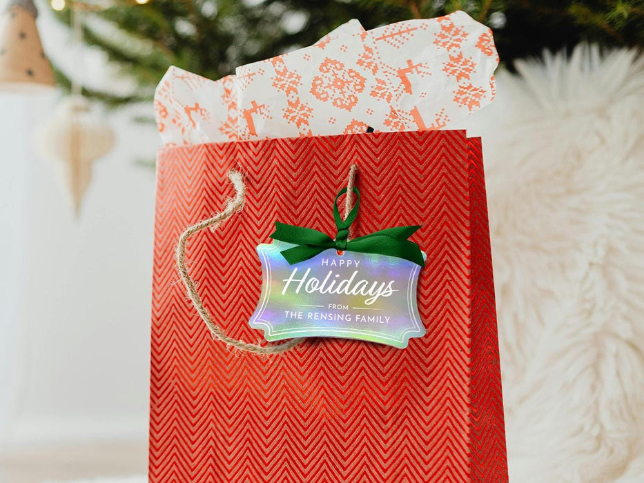 A iridescent cardstock Happy Holidays gift tag is shown hanging from red gift bag. The bag sits under a Christmas tree against a white background.