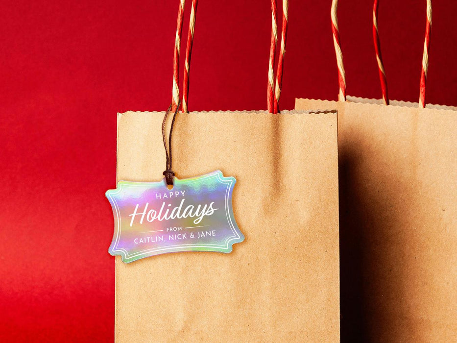 An iridescent cardstock Happy Holidays gift tag is shown hanging from a brown, craft paper gift bag. The bag is on a red backdrop.