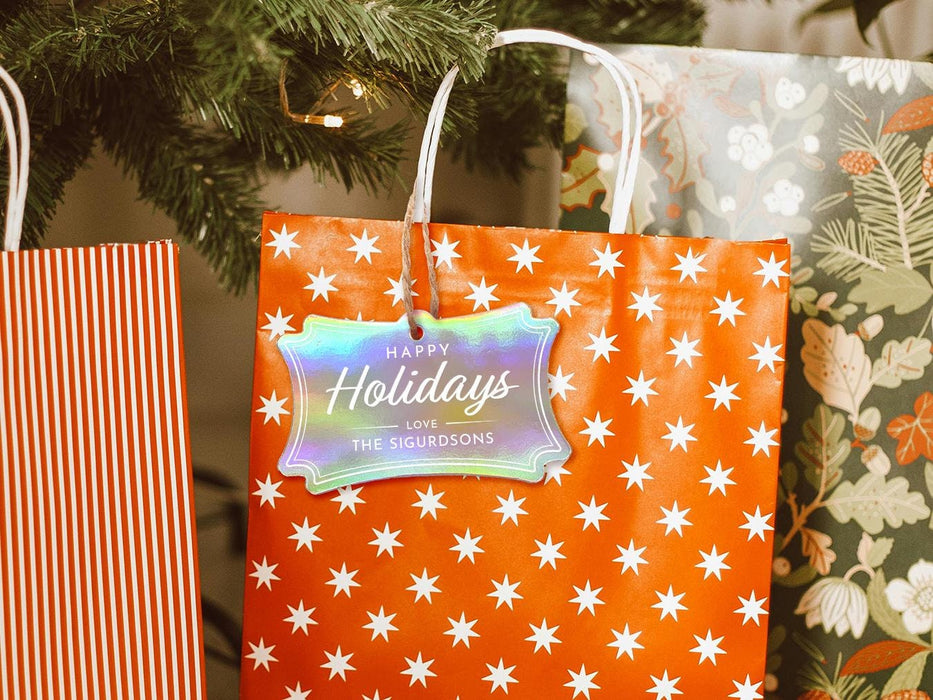 An iridescent cardstock Happy Holidays gift tag is shown hanging from a star-patterned white and red gift bag. The bag sits under a Christmas tree surrounded by other presents.