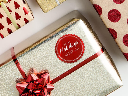 A red foil cardstock Happy Holidays gift tag is shown on a Christmas present. The present is wrapped in gold paper and has a red bow and ribbon around it. The background of the picture is a white surface.