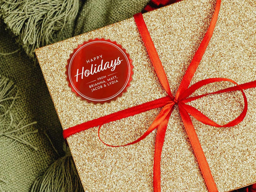 A red foil cardstock Happy Holidays gift tag is shown on a glitter wrapped box. The box also has a red ribbon on it. The box is seen on a green blanket surface.