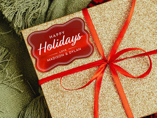 A red foil cardstock Happy Holidays gift tag is shown on a glitter wrapped box. The box also has a red ribbon on it. The box is seen on a green blanket surface.