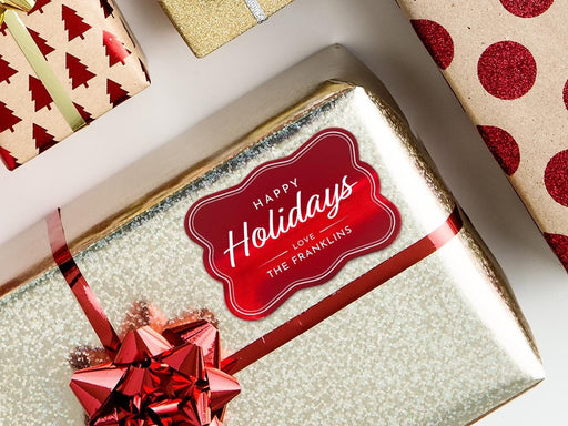 A red foil cardstock Happy Holidays gift tag is shown on a Christmas present. The present is wrapped in gold paper and has a red bow and ribbon around it. The background of the picture is a white surface.