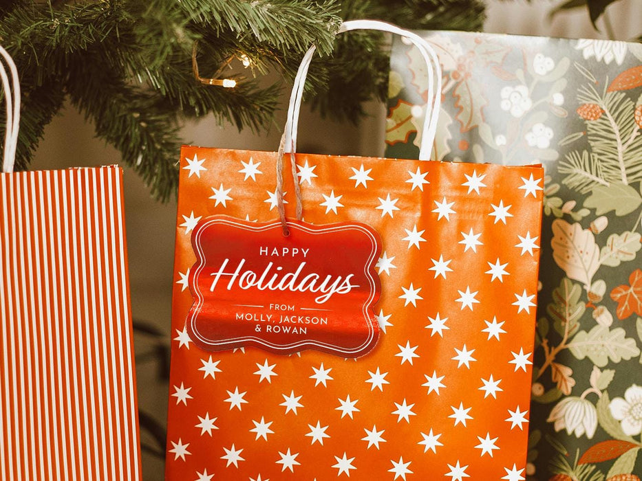 A red foil cardstock Happy Holidays gift tag is shown hanging from a star-patterned white and red gift bag. The bag sits under a Christmas tree surrounded by other presents.