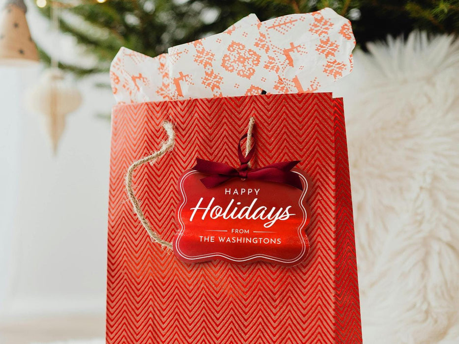 A red foil cardstock Happy Holidays gift tag is shown hanging from red gift bag. The bag sits under a Christmas tree against a white background.