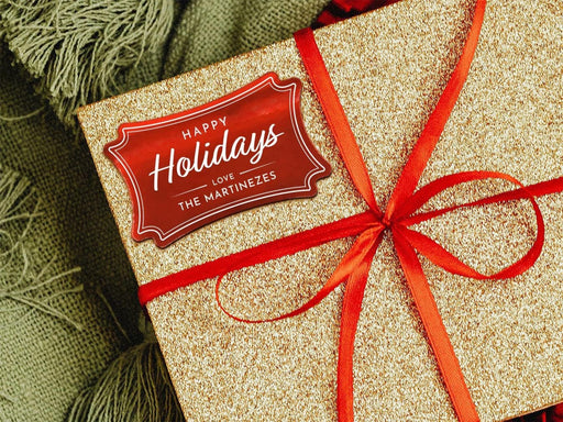 A red foil cardstock Happy Holidays gift tag is shown on a glitter wrapped box. The box also has a red ribbon on it. The box is seen on a green blanket surface.