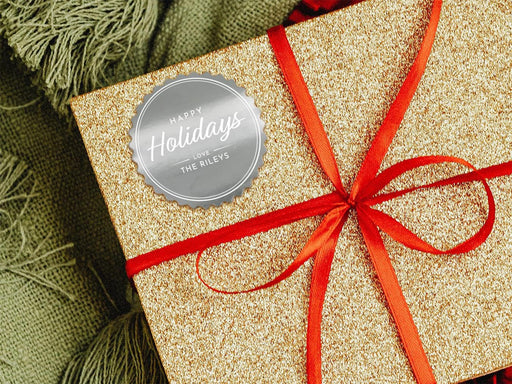 A silver foil cardstock Happy Holidays gift tag is shown on a glitter wrapped box. The box also has a red ribbon on it. The box is seen on a green blanket surface.