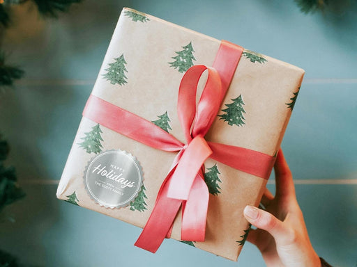 A silver foil cardstock Happy Holidays gift tag is shown on a Christmas present. The present is wrapped in Christmas tree paper and has a red ribbon around it. The background of the picture is a green/blue.