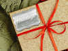 A silver foil cardstock Happy Holidays gift tag is shown on a glitter wrapped box. The box also has a red ribbon on it. The box is seen on a green blanket surface.