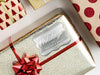 A silver foil cardstock Happy Holidays gift tag is shown on a Christmas present. The present is wrapped in gold paper and has a red bow and ribbon around it. The background of the picture is a white surface.