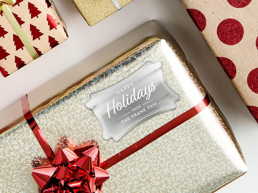 A silver foil cardstock Happy Holidays gift tag is shown on a Christmas present. The present is wrapped in gold paper and has a red bow and ribbon around it. The background of the picture is a white surface.