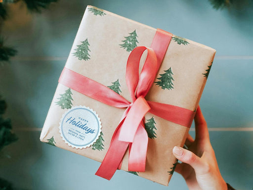 A white glitter cardstock gift tag printed with blue ink is shown on a Christmas present. The present is wrapped in Christmas tree paper and has a red ribbon around it. The background of the picture is a green/blue.