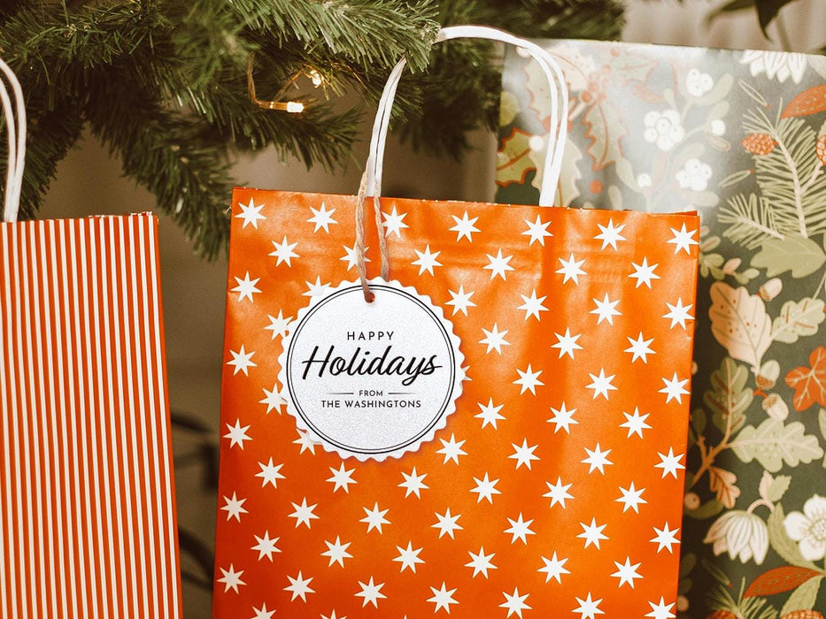 A silver foil cardstock Happy Holidays gift tag with black ink is shown hanging from a star-patterned white and red gift bag. The bag sits under a Christmas tree surrounded by other presents.