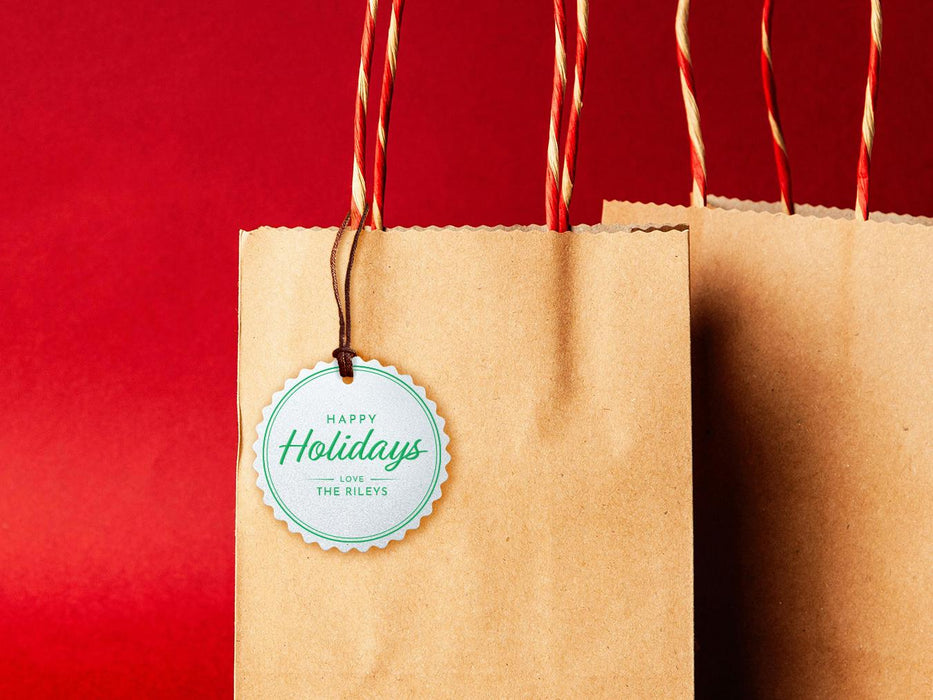 A white glitter cardstock Happy Holidays gift tag with green ink is shown hanging from a brown, craft paper gift bag. The bag is on a red backdrop.