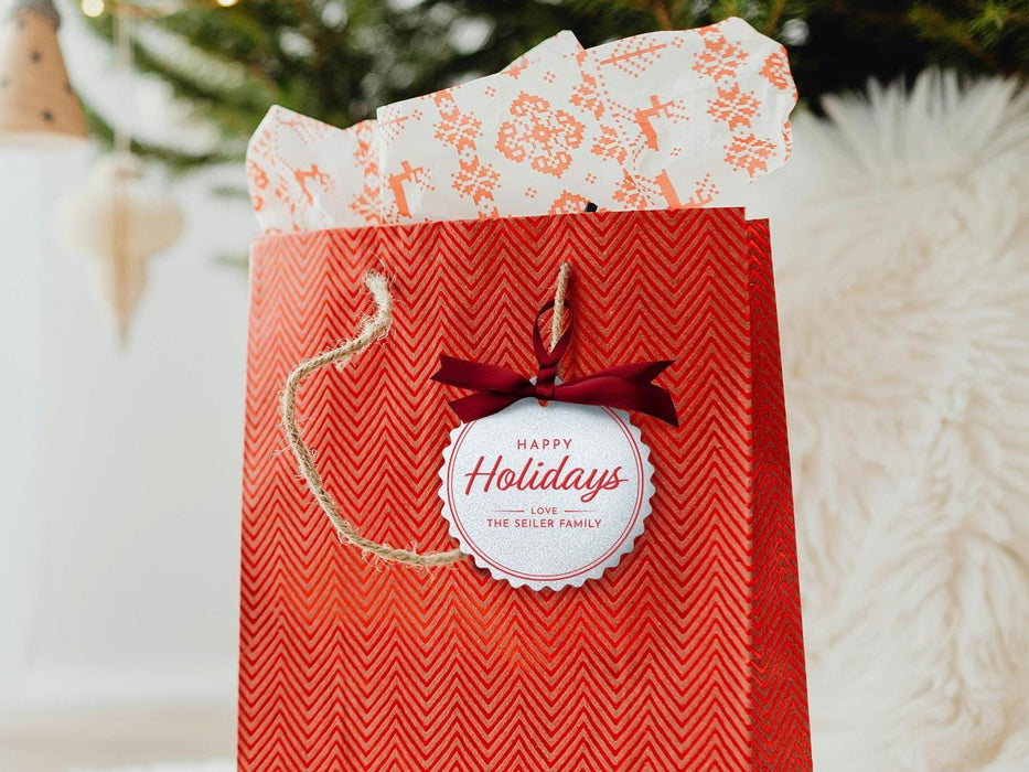 A white glitter cardstock Happy Holidays gift tag with red ink is shown hanging from red gift bag. The bag sits under a Christmas tree against a white background.