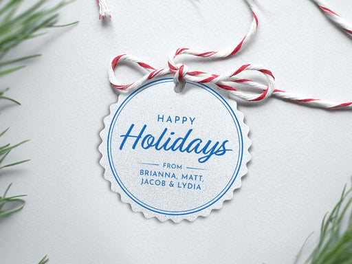 A silver foil cardstock Happy Holidays gift tag with blue ink is shown sitting on a white background surrounded by Christmas tree clippings. The tag has a red and white string looped through the top of it.