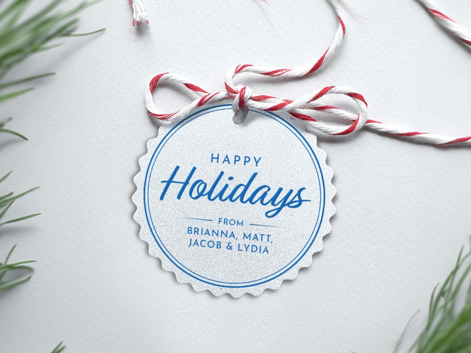 A silver foil cardstock Happy Holidays gift tag with blue ink is shown sitting on a white background surrounded by Christmas tree clippings. The tag has a red and white string looped through the top of it.