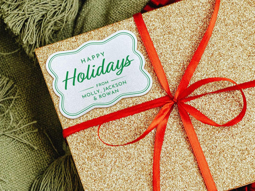 A white glitter cardstock Happy Holidays gift tag with green ink is shown on a glitter wrapped box. The box also has a red ribbon on it. The box is seen on a green blanket surface.