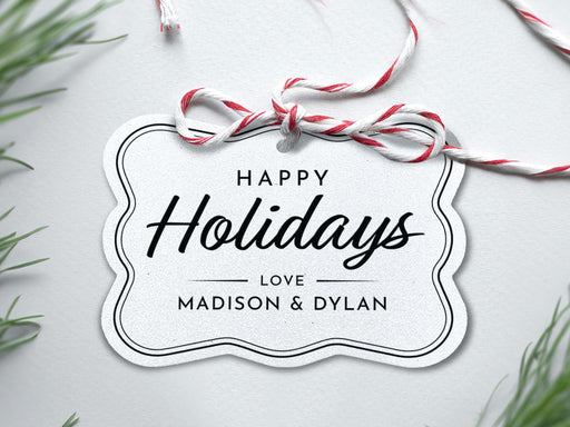 A silver foil cardstock Happy Holidays gift tag with black ink is shown sitting on a white background surrounded by Christmas tree clippings. The tag has a red and white string looped through the top of it.