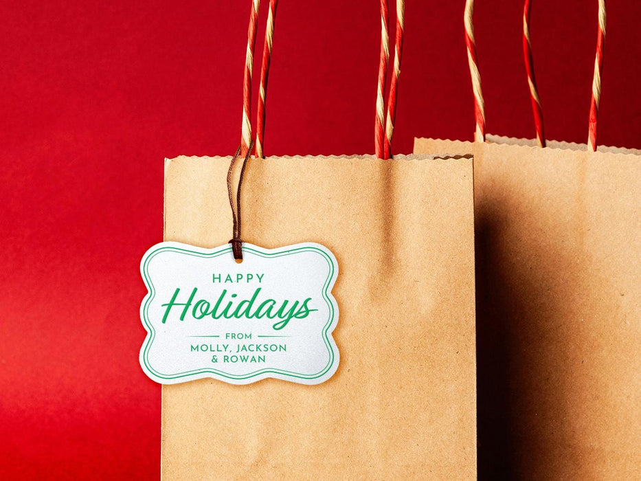 A white glitter cardstock Happy Holidays gift tag with green ink is shown hanging from a brown, craft paper gift bag. The bag is on a red backdrop.