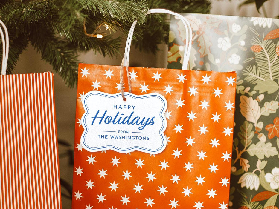 A silver foil cardstock Happy Holidays gift tag with blue ink is shown hanging from a star-patterned white and red gift bag. The bag sits under a Christmas tree surrounded by other presents.