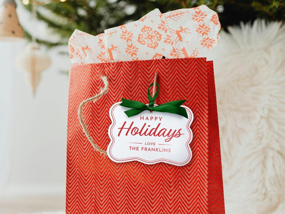 A white glitter cardstock Happy Holidays gift tag with red ink is shown hanging from red gift bag. The bag sits under a Christmas tree against a white background.