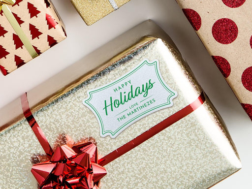 A white glitter cardstock Happy Holidays gift tag with green ink is shown on a Christmas present. The present is wrapped in gold paper and has a red bow and ribbon around it. The background of the picture is a white surface.