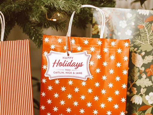 A silver foil cardstock Happy Holidays gift tag with red ink is shown hanging from a star-patterned white and red gift bag. The bag sits under a Christmas tree surrounded by other presents.