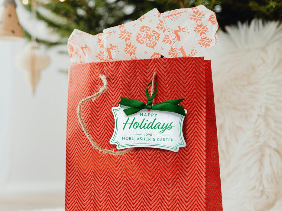 A white glitter cardstock Happy Holidays gift tag with green ink is shown hanging from red gift bag. The bag sits under a Christmas tree against a white background.