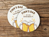 Two coasters sit on a wood background. Coasters feature bar design. This design uses brown and orange lettering, and a drawing of beer glasses. The name, established date, and location can be customized.