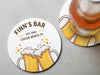 Two coasters sit on a grey surface with a beer on top of one. Coasters feature bar design. This design uses brown and orange lettering, and a drawing of beer glasses. The name, established date, and location can be customized.