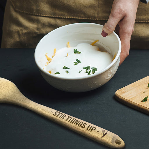 Wooden spoon with an engraving on the handle that says Stir Things Up with whisk and pin icon, on black table next to mixing bowl and cutting board with herbs on it.