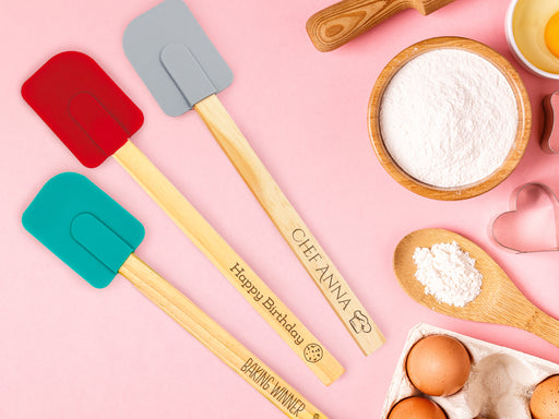 three silicone spatulas with wooden handles with engravings on handles that say Baking Winner With whisk icon, Happy Birthday with cookie icon, and Chef Anna with chef hat icon. Spatulas are on pink background surrounded by baking supplies