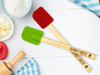 two custom engraved wooden spatulas on a white wooden table surrounded by baking supplies