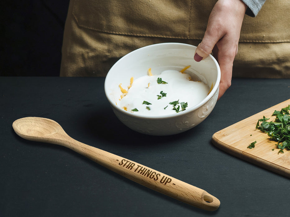 wooden spoon with engraving on handle that says Stir Things up on back table in front of person wearing apron making a sauce in a bowl next to a cutting board