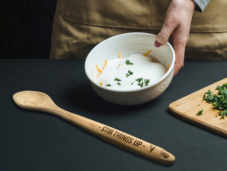 Wooden spoon on black table next to mixing bowl and cutting board with herbs on it.