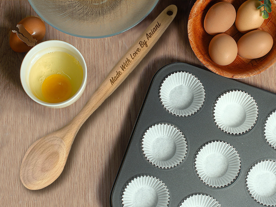 wooden spoon on wooden table surrounded by kitchen supplies such as pans, bowls, eggs, on the handle is engraved Made with Love By Ariani