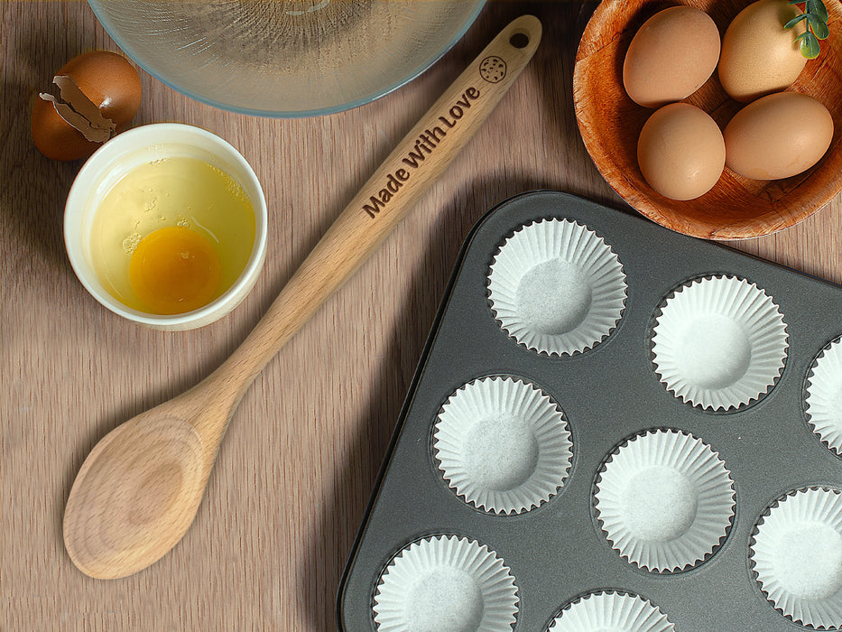 wooden spoon on wooden table surrounded by kitchen supplies such as pans, bowls, eggs, on the handle is engraved Made with Love with cookie icon.