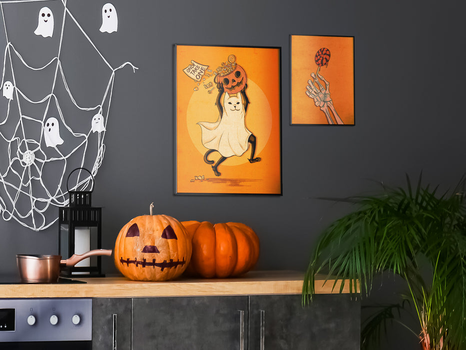 2 frames of wall art with halloween designs on a kitchen wall with a countertop with pumpkins and spider ghost decoration. art designs include; a cat dressed as a ghost carrying a pumpkin, and a skeleton hand