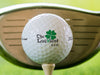 single white titleist golf ball with custom The Luckiest Design on beige tee in front of golf club with grass background